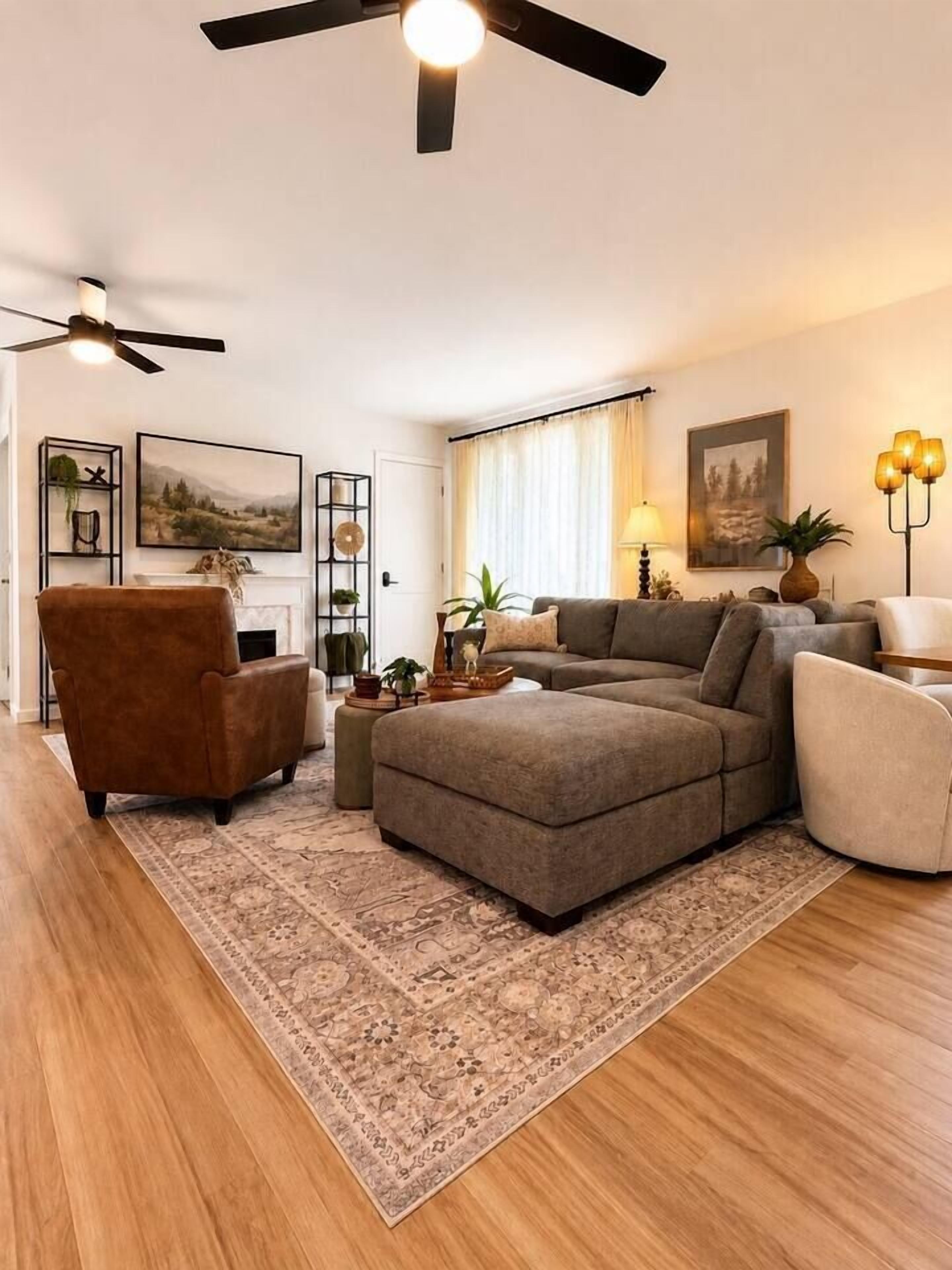 A living room with a sectional sofa, a brown armchair, a white armchair, a fireplace, and a ceiling fan.