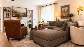 A living room with a sectional sofa, a brown armchair, a white armchair, a fireplace, and a ceiling fan.