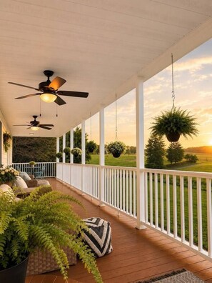 A covered porch with ceiling fans, hanging planters, and a view of a grassy landscape.
