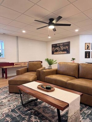 A room with a brown leather sofa, a wooden coffee table, a ceiling fan, and a pool table.