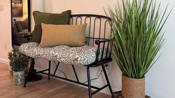 Una habitación con una silla de estructura metálica y cojines estampados, una planta grande en maceta, un cuadro en la pared y un espejo.