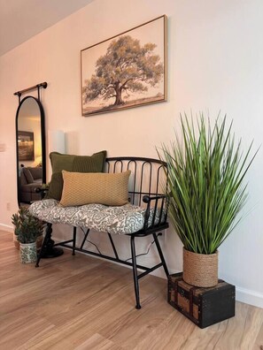 Ein Zimmer mit einem stahlrohrgestützten Stuhl mit gemusterten Kissen, einer großen Topfpflanze, einem gerahmten Gemälde an der Wand und einem Spiegel.