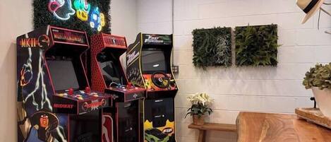 Una habitación con tres máquinas de videojuegos, una mesa de madera y una planta en la pared.