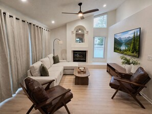 A modern living room with a sectional sofa, two leather chairs, a coffee table, a fireplace, a ceiling fan, and a large TV mounted on the wall. - Sand and Sage: 3BR, 2BH, Heated Pool, Fenced Yard (Spring Hill)
