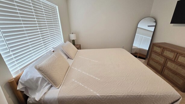 Een net opgemaakt bed met een witte deken en kussens, een houten nachtkastje met een lamp, een houten dressoir en een grote spiegel aan de muur.