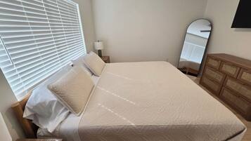 A neatly made bed with a white comforter and pillows, a wooden nightstand with a lamp, a wooden dresser, and a large mirror on the wall.