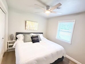 Une chambre à coucher comprenant un lit, un ventilateur de plafond, une fenêtre avec des stores et une œuvre d’art accrochée au mur.
