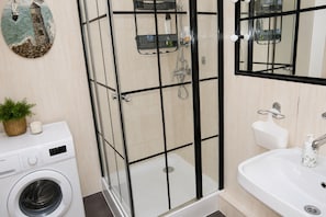 A bathroom with a glass-enclosed shower, a washing machine, a sink, and a decorative wall piece. - Mrzezyno Holiday Home for 5 People (Trzebiatow)