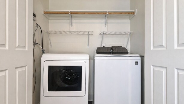 A laundry room with a front-loading washer and dryer, a shelf above the dryer, and a wall-mounted electrical outlet. - Pet Friendly, Short Drive to Beach, Nearby Stores! (Galveston)