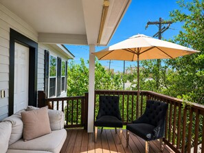 A covered porch with a wooden deck, a beige sofa with cushions, two black armchairs, and a large beige umbrella providing shade. - Pet Friendly, Short Drive to Beach, Nearby Stores! (Galveston)