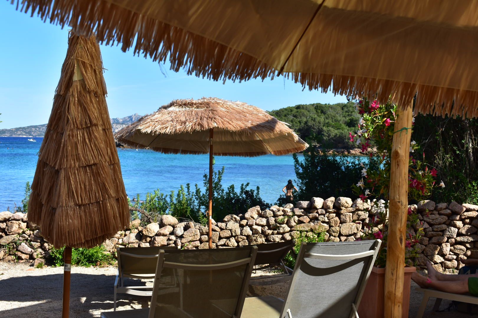 Une terrasse en bord de mer, agrémentée de parasols en chaume, de murs en pierre et de sièges d’extérieur.