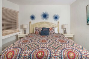 Ein Schlafzimmer mit einem Musterbettüberwurf, einem Holzkopfende, zwei Nachttischlampen und einem Fenster mit Jalousien.