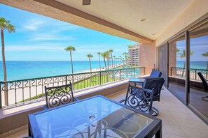 Ein Balkon mit Glastisch und Stühlen, der Blick auf einen Strand mit Palmen und einem wolkenlosen, blauen Himmel bietet.