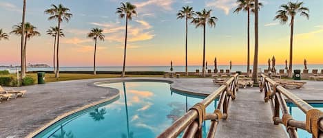 Zona de piscina con palmeras y vista al océano al atardecer.