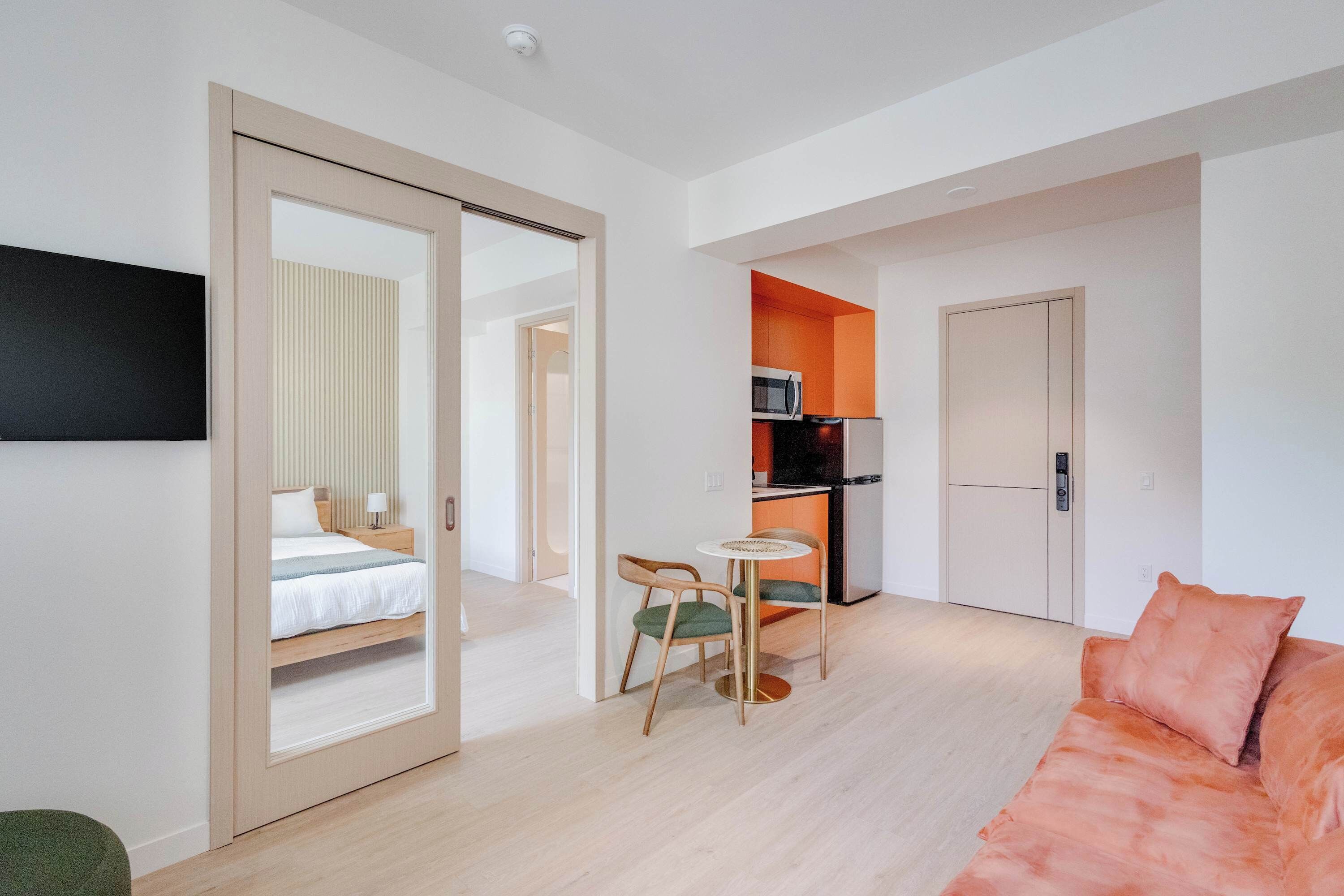 A modern hotel room with a bed, a small dining area with a table and chairs, a microwave, and a refrigerator.