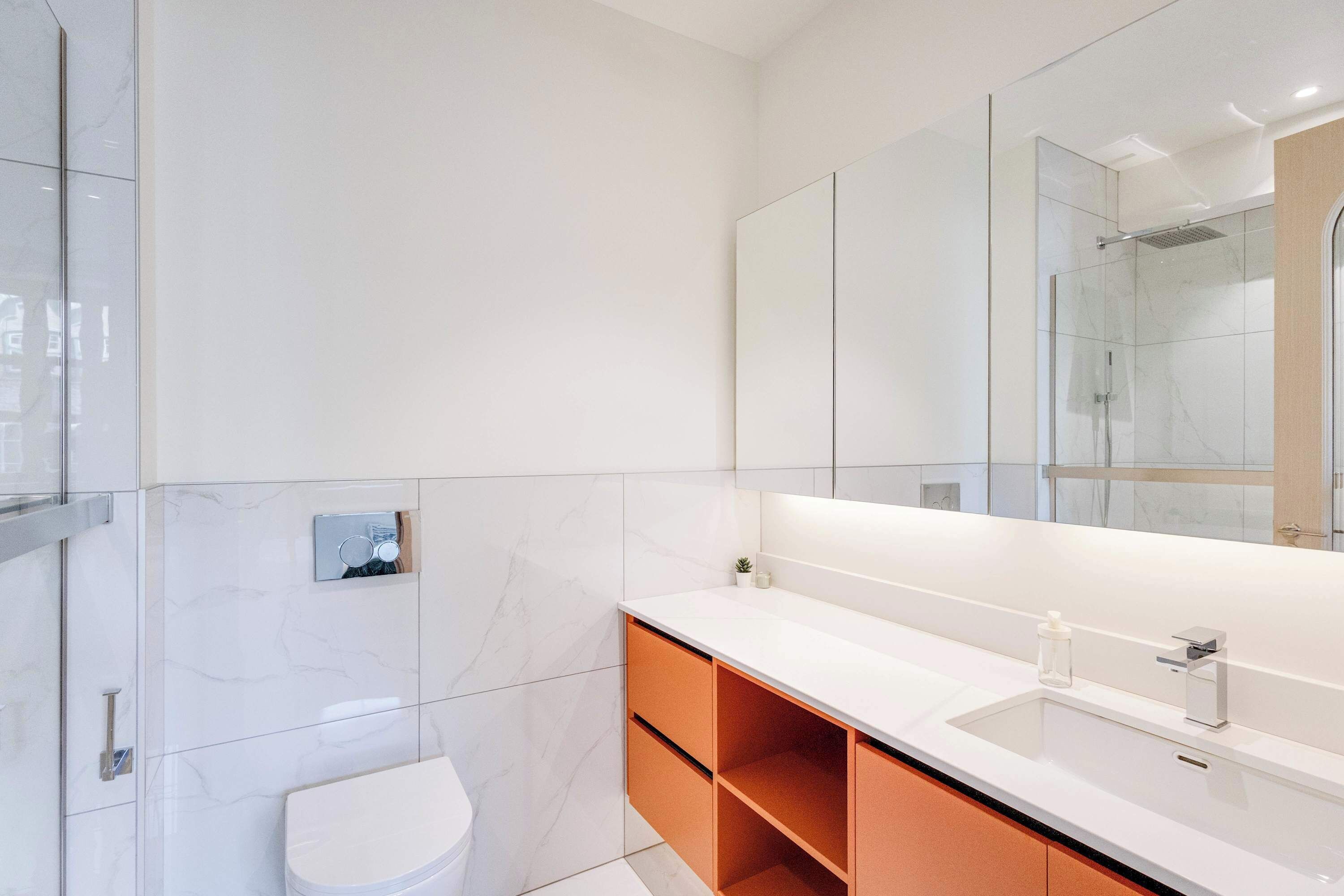 A modern bathroom with a white toilet, a glass shower enclosure, and a large mirror above a white sink with a wooden cabinet below.