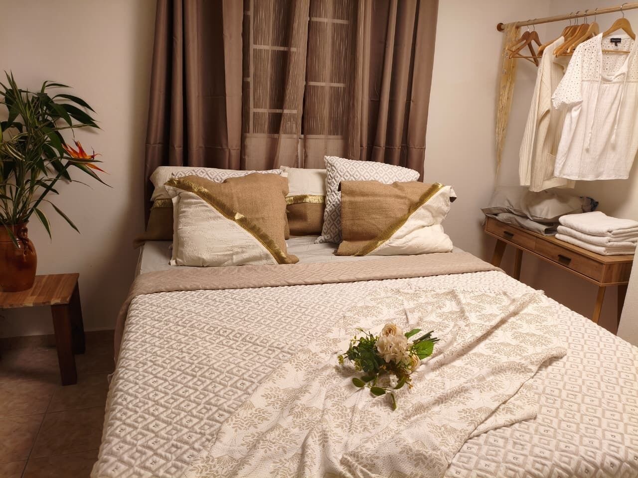 Un lit bien fait, avec une literie à motifs, une table de chevet ornée d’une plante et un portant à vêtements sur lequel sont pliés des vêtements.