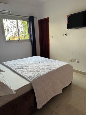 A bedroom with a bed, a window with decorative iron bars, a dark curtain, a wooden door, and a mounted television.