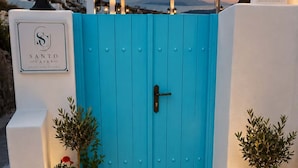 Ein blaues Tor mit Schlüsselloch, flankiert von Kübelpflanzen mit roten Blüten, und ein Schild für 'SANTO CAVES' an einer weißen Mauer.