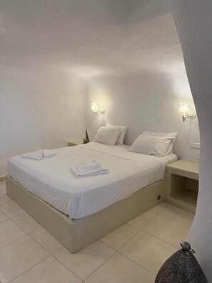 A neatly made bed with white linens and towels, a bedside table with a lamp, and a nightstand with a small decorative item. - Santo Caves Santorini Suite With Private Hot Tub (Santorini)