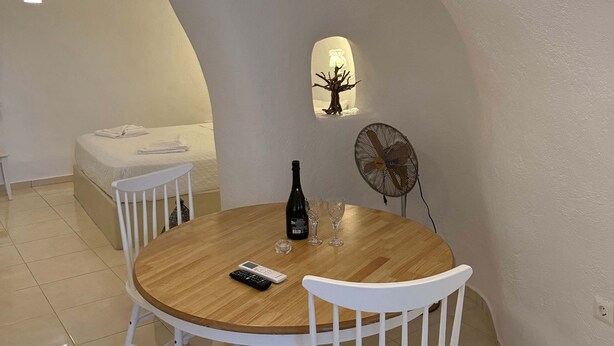 A dining area with a round wooden table, white chairs, a bottle of wine, and glasses. - Santo Caves Santorini Suite With Private Hot Tub (Santorini)