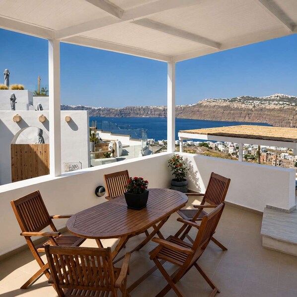 A balcony with a wooden table and chairs, a potted plant, and a view of the sea and buildings. - Santo Cave Santorini Suite With Seaviews (Santorini)