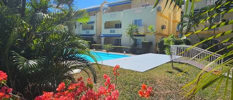 A pool area with a clear blue sky, surrounded by lush greenery and red flowers.