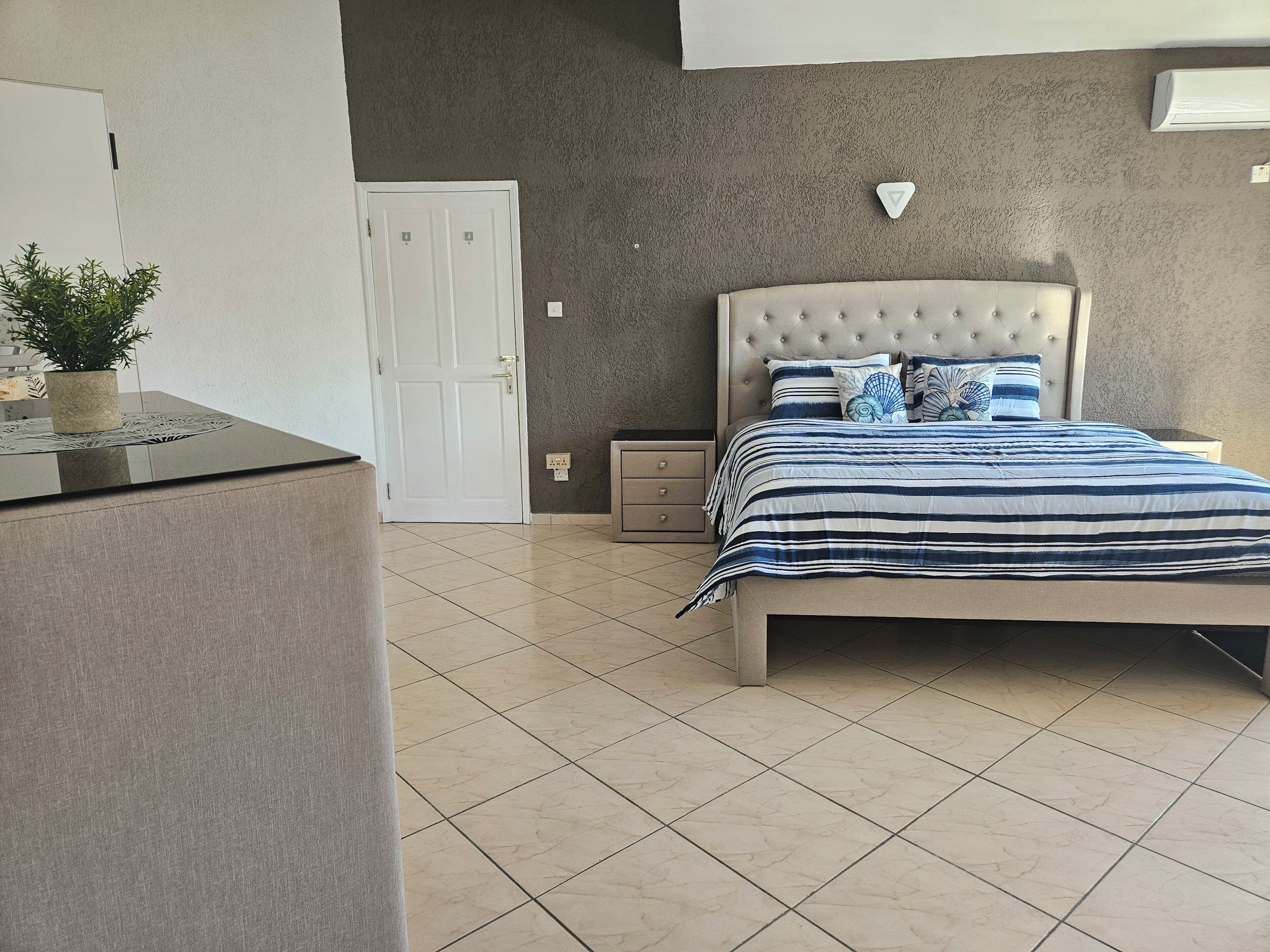 Une chambre à coucher avec un lit, des tables de chevet, une plante en pot et une porte.