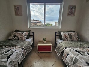 A bedroom with two beds, a small table, and a window with a view of buildings. - Le Coin Grenadier (Flic en Flac)