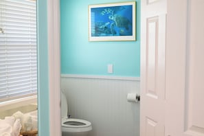 A bathroom with a toilet, a framed picture of a sea turtle, and a window with blinds. - Stylish Deluxe Gulf View studio, with jacuzzi tub (Bradenton Beach)