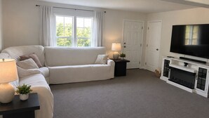 Ett vardagsrum med en vit soffa, en tv, en öppen spis och ett fönster med gardiner.
