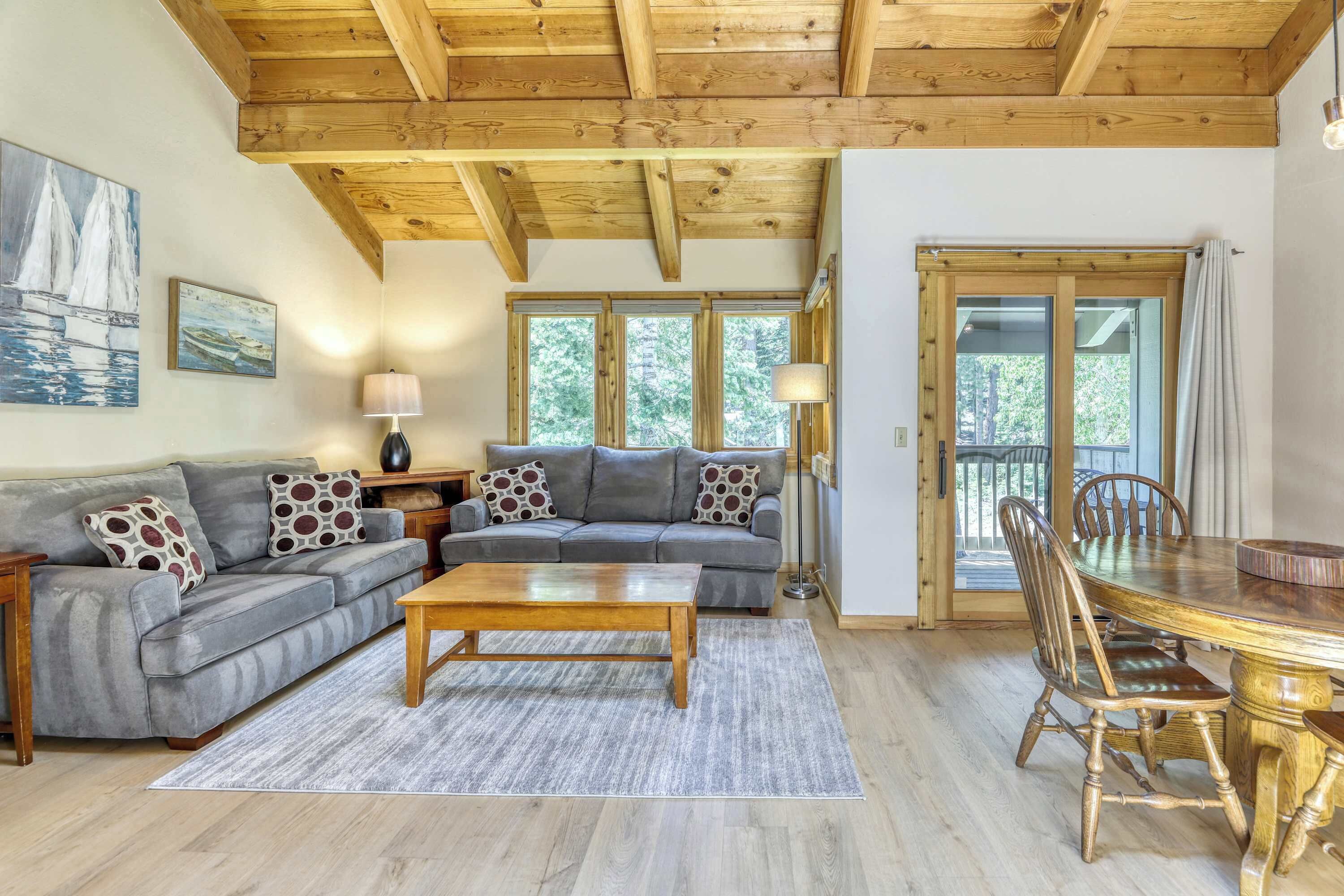 A living room with a grey sofa, wooden coffee table, and dining table with chairs. There are windows and a door leading outside.