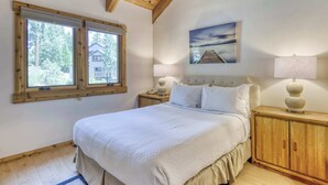 A bedroom with a large bed, wooden headboard, a nightstand with a lamp, a window with a view of a house, and a painting on the wall. - Condo at Granlibakken Tahoe Resort - Ski-In - Ski-Out with Pool, Spa & Minutes from Lake Tahoe (Tahoe City)