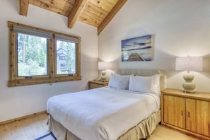 A bedroom with a large bed, wooden headboard, a nightstand with a lamp, a window with a view of a house, and a painting on the wall. - Condo at Granlibakken Tahoe Resort - Ski-In - Ski-Out with Pool, Spa & Minutes from Lake Tahoe (Tahoe City)