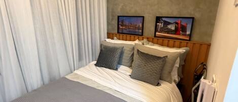 Une chambre d’hôtel avec un lit, une tête de lit, une fenêtre avec des rideaux et un tableau accroché au mur.