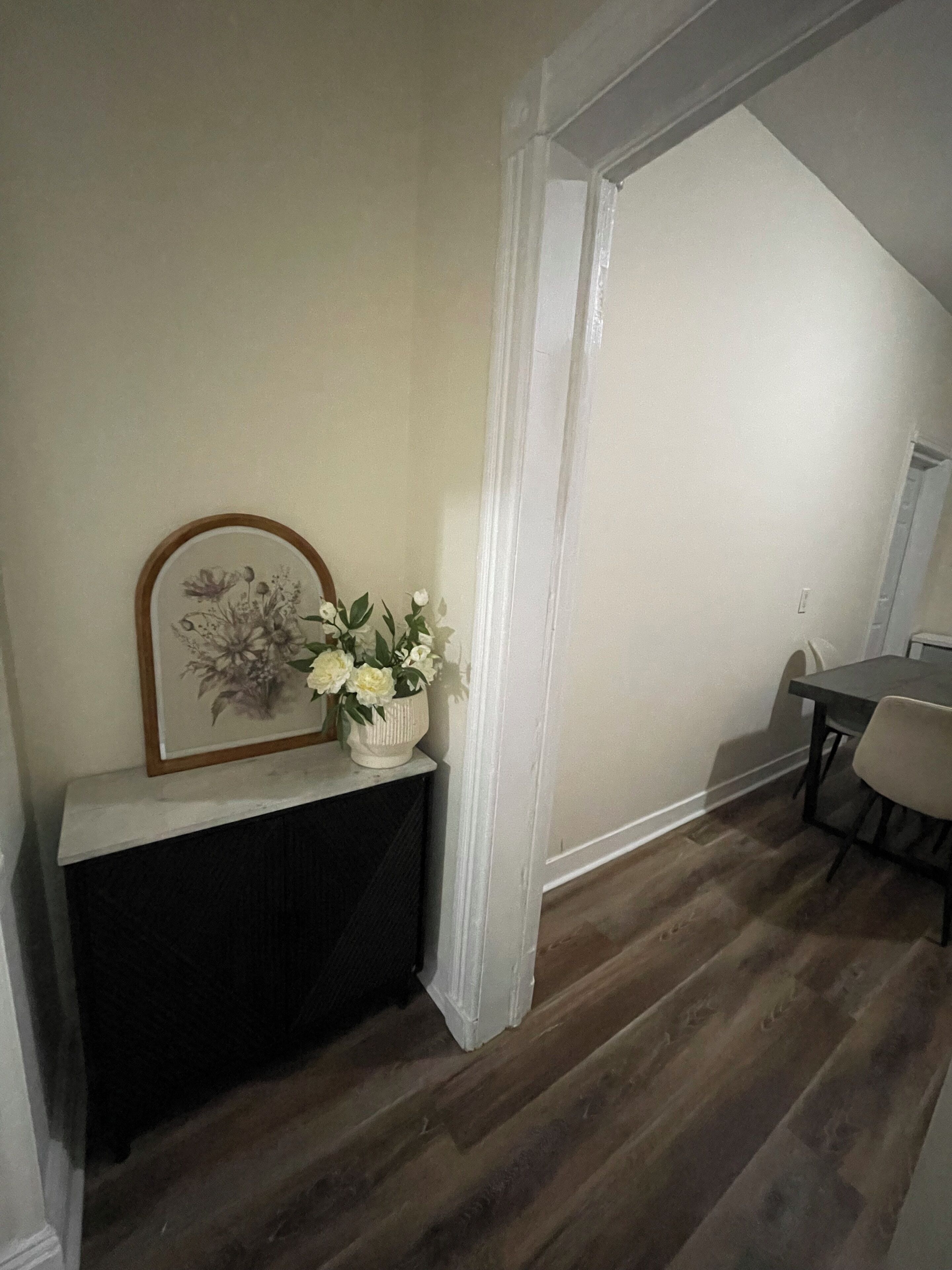 Ein Zimmer mit dunklem Holzboden, einem weißen Konsolentisch mit einem gerahmten Blumenbild und einer weißen Vase mit Blumen.