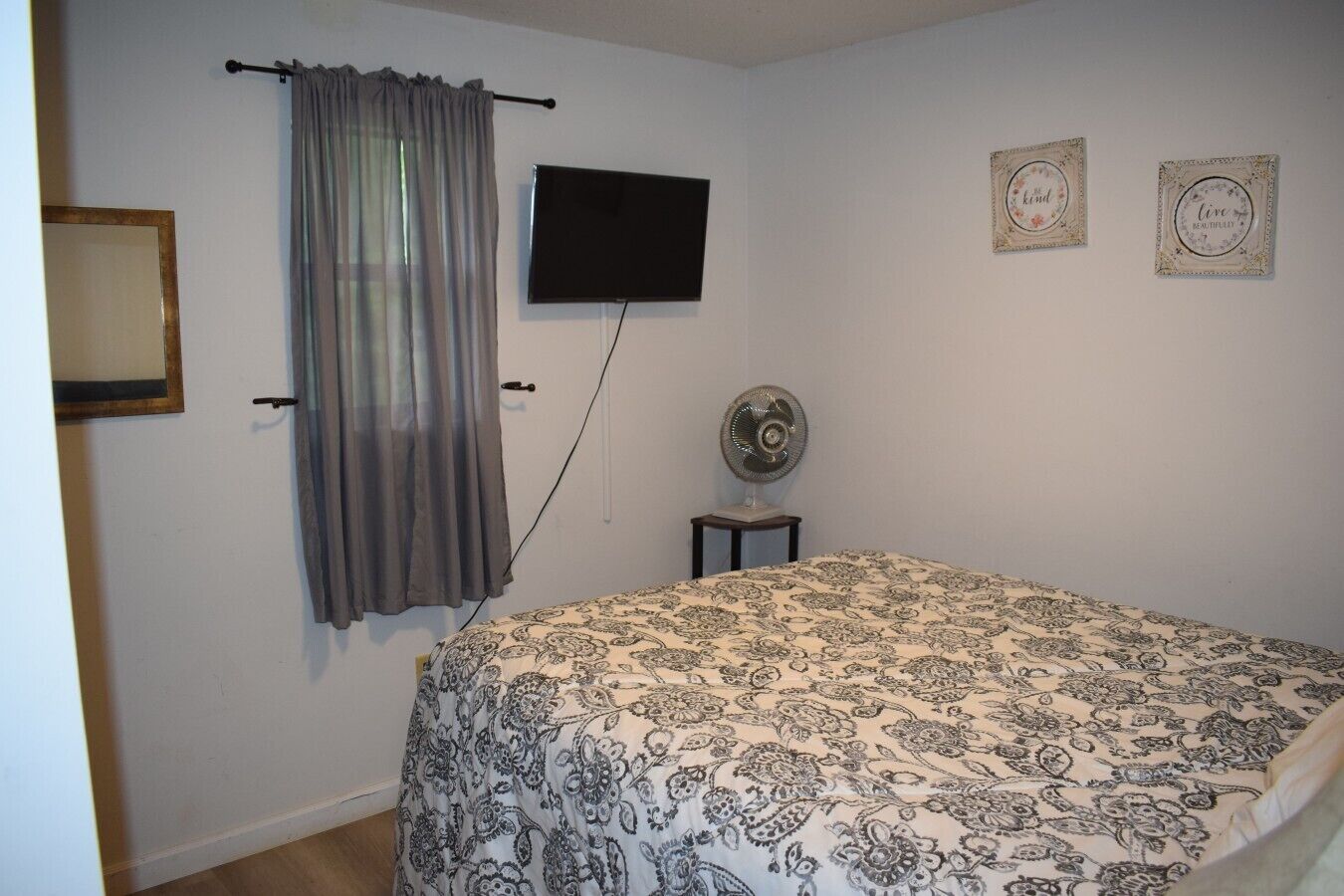 Een slaapkamer met een bed, een tv aan de muur, een ventilator op een tafeltje en een gordijn.