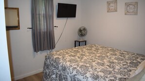 Een slaapkamer met een bed, een tv aan de muur, een ventilator op een tafeltje en een gordijn.