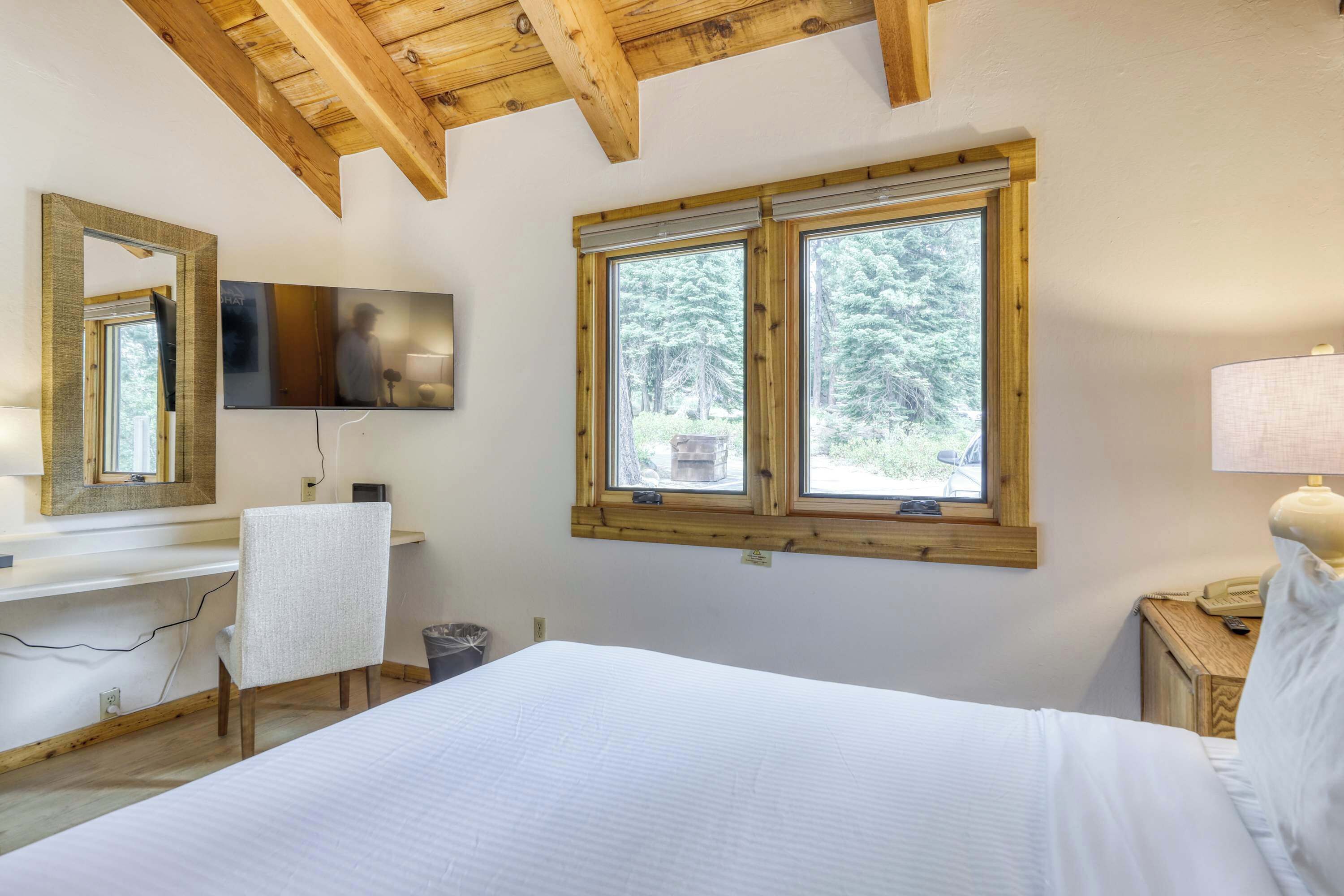 A bedroom with a bed, a desk, a chair, a TV mounted on the wall, and a window with a view of trees.