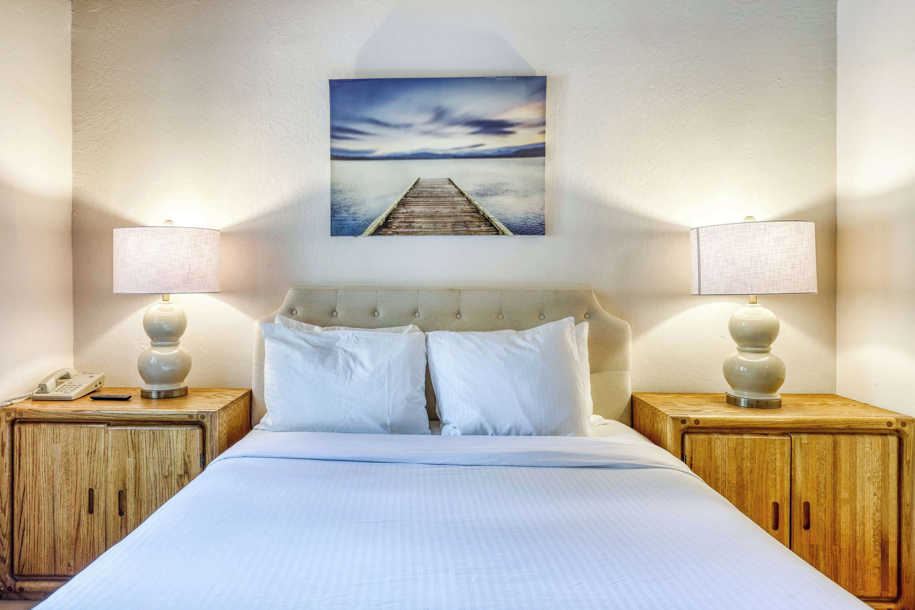 Habitación de hotel con cama, dos lámparas de noche, un teléfono y un cuadro de un muelle.