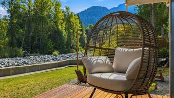 Une chaise en osier avec des coussins, placée sur une terrasse en bois, d’où l’on peut admirer une rive rocheuse et des montagnes boisées.