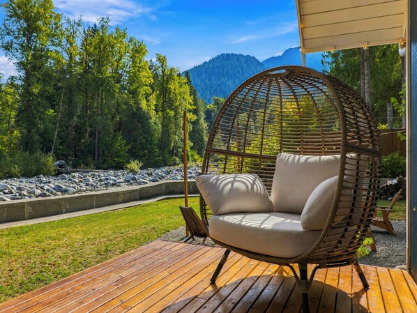 A wicker chair with cushions on a wooden deck, overlooking a rocky riverbank and forested mountains.
