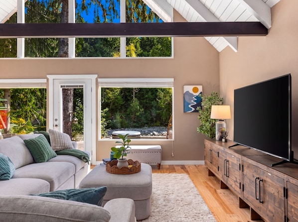 A modern living room with a sofa, ottoman, and TV stand, large windows with a view of trees, and a ceiling with exposed beams.