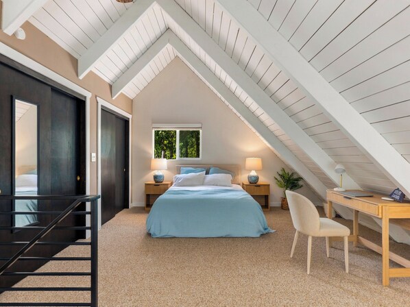 Une chambre avec un plafond en pente, un lit avec une literie bleue, deux tables de chevet avec des lampes, une chaise et un bureau avec un ordinateur.