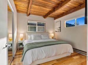 A bedroom with a wooden ceiling, a bed with white bedding, two bedside lamps, a window with blinds, and a framed picture on the wall. - Riverfront A-Frame — Hot Tub, Sauna & Steam Shower (Granite Falls)