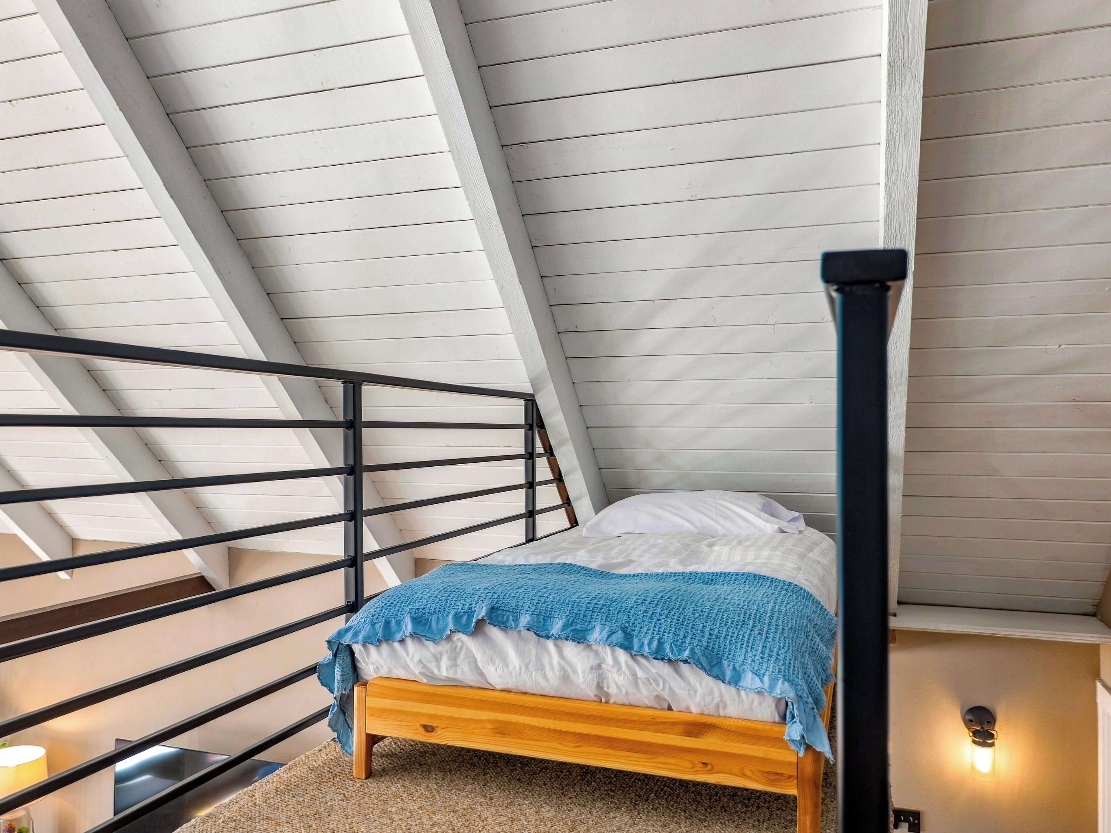 A bedroom with a sloped ceiling, a wooden bed, a black metal railing, and a wall-mounted light fixture.