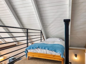 A bedroom with a sloped ceiling, a wooden bed, a black metal railing, and a wall-mounted light fixture.