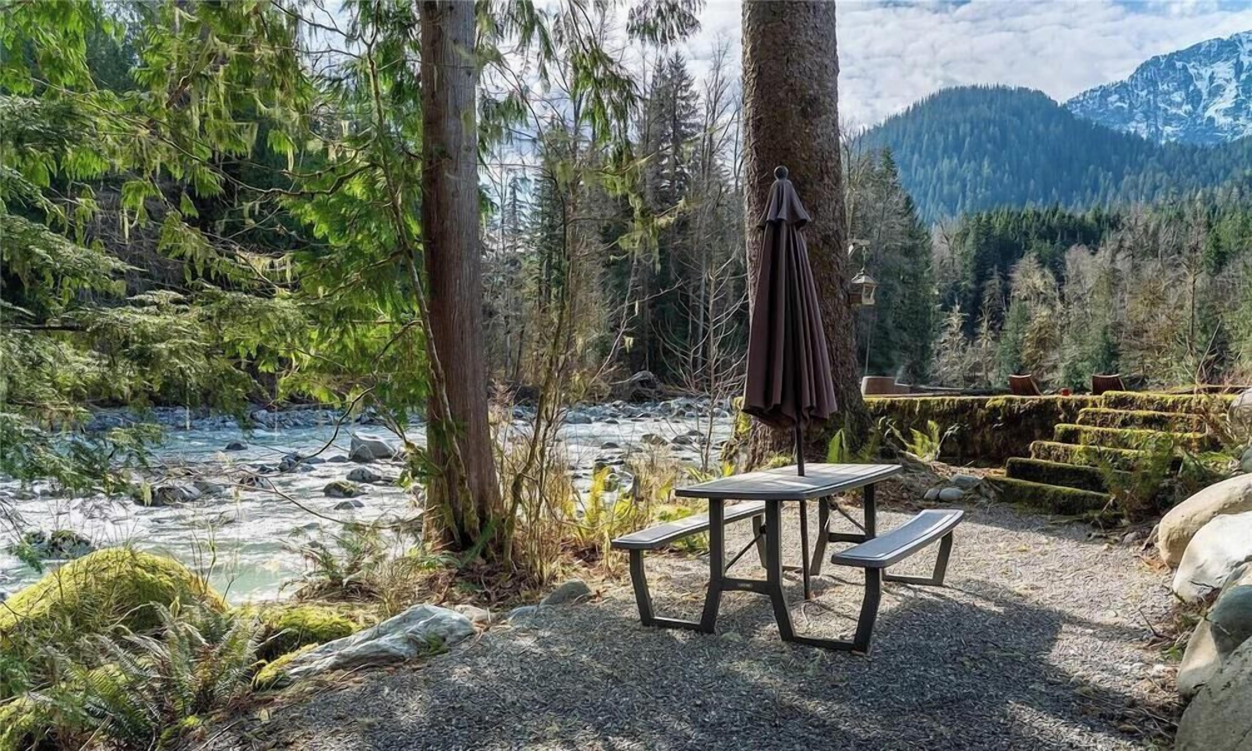 Ein Picknicktisch mit Bänken und einem hängenden Sonnenschirm in einer bewaldeten Gegend, im Hintergrund ein Fluss und Berge.
