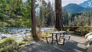 Une table de pique-nique avec des bancs et un parasol suspendu, située dans une zone boisée, avec une rivière et des montagnes en arrière-plan.