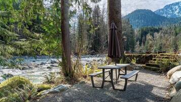 Ein Picknicktisch mit Bänken und einem hängenden Sonnenschirm in einer bewaldeten Gegend, im Hintergrund ein Fluss und Berge.